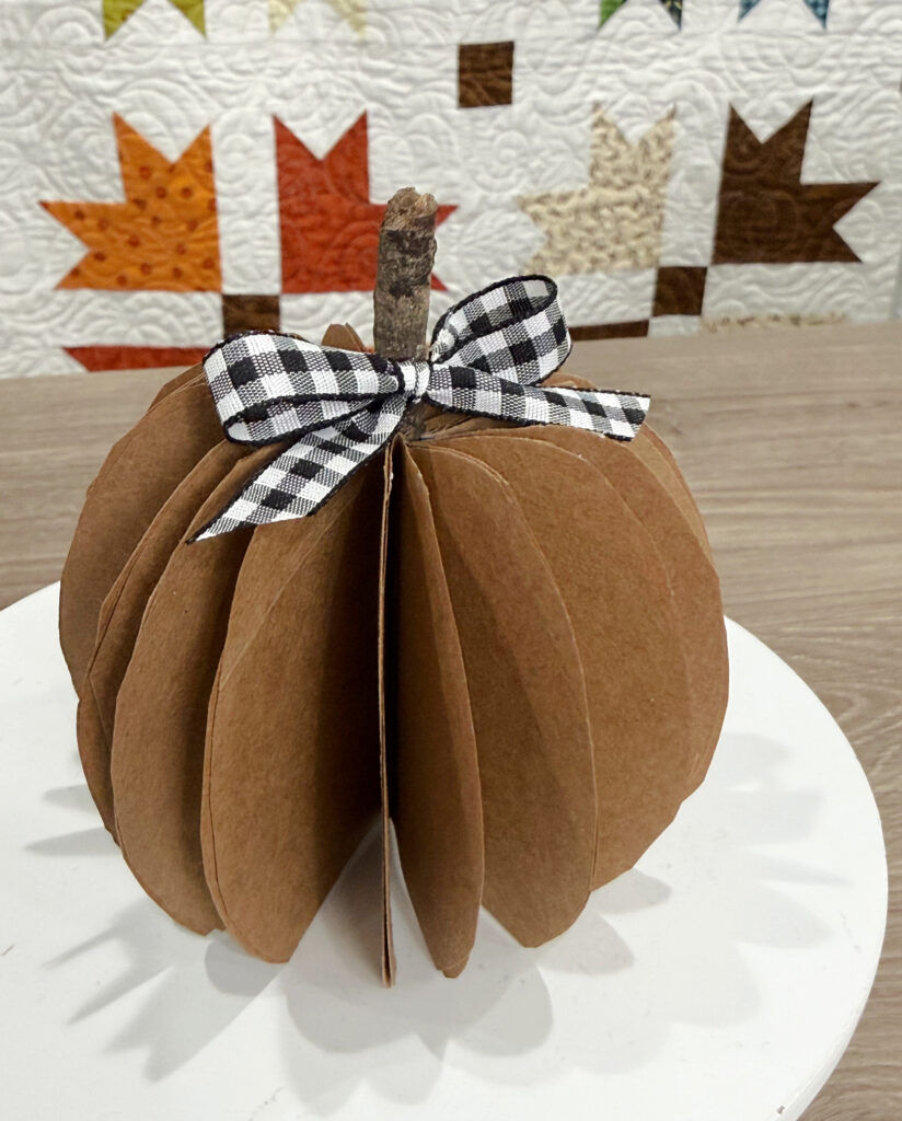 Image contains a paper bag pumpkin on a white tray sitting in front of an autumn quilt.