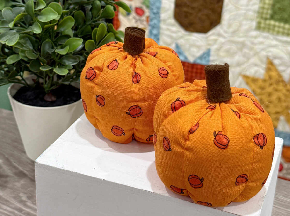 Image contains two completed fabric pumpkins on a white box next to a faux plant.