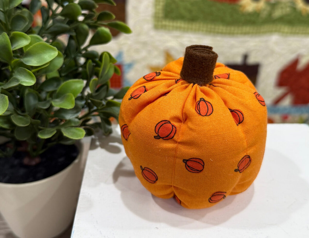 Image contains a finished fabric pumpkin on a white box next to a faux plant.